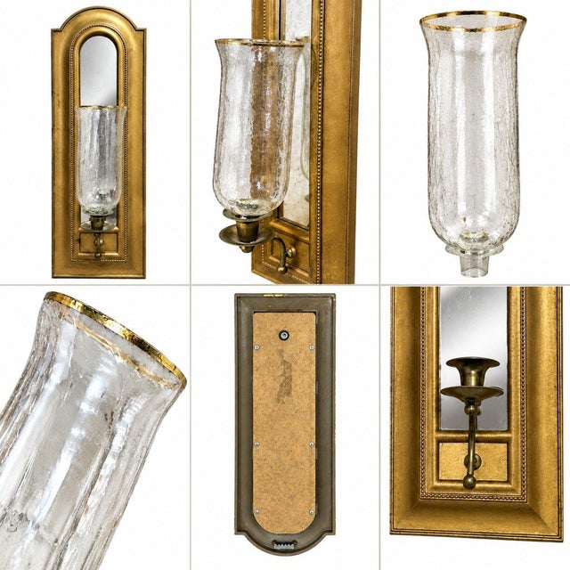 An elegant antique wall holder featuring a tall arched brass frame with a built-in mirror and a delicate glass hurricane...