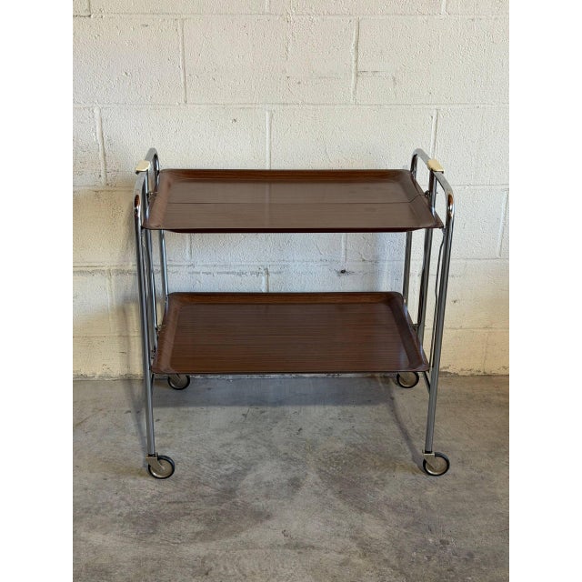 This two-tier folding bar cart features rich wood surfaces supported by a sleek metallic frame on smooth-rolling casters....