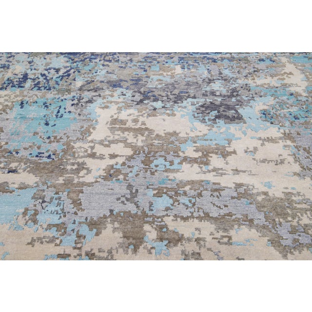 This Indian wool and silk blend rug features a light gray field with an abstract pattern detailing blue, brown, and beige...