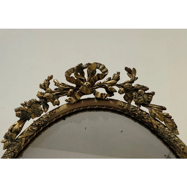Antique oval mirror in gilt bronze, featuring a finely detailed floral and bow crest. The frame showcases classical...