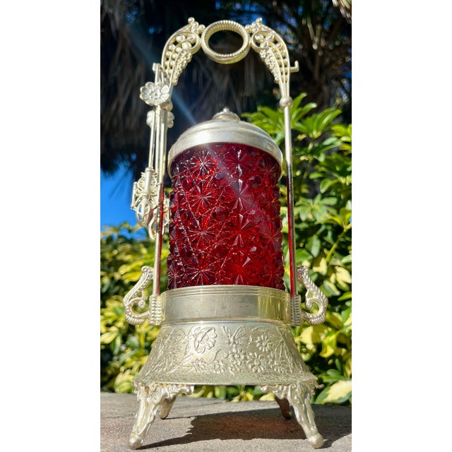 Decorative silver plated castor with lid and tongs. Features a deep ruby red glass jar in daisy and button pattern.