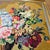 Hand-stitched needlepoint tapestry features a vibrant floral still life arrangement in a classical urn, set against a rich...