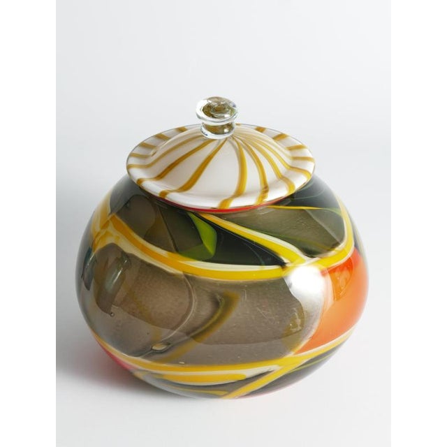 This vibrant, one-of-a-kind lidded vessel features a bulbous, spherical body with a translucent, smoky-grey base that is...