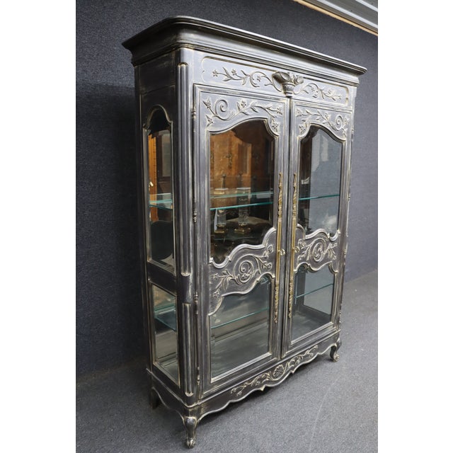 French Provincial–style display cabinet with carved floral and scroll details and a distressed charcoal finish. The piece...