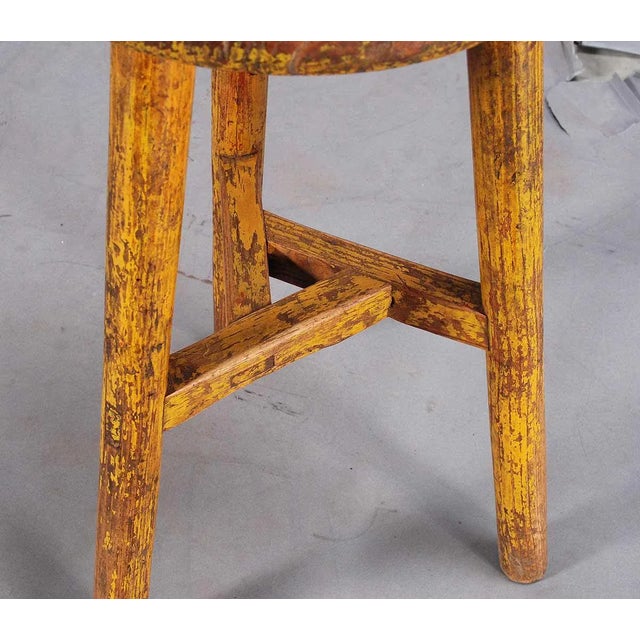 A three-legged elm stool with distressed paint finish. The turned legs extend through the seat and are joined by stretchers.