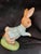 A ceramic figurine of Peter Rabbit in a blue jacket, running on a green base. The figurine is part of a collection...