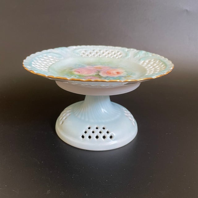 A lovely stemmed porcelain candy dish from the 1900s. Hand painted roses and small purple flowers in the center, accented...