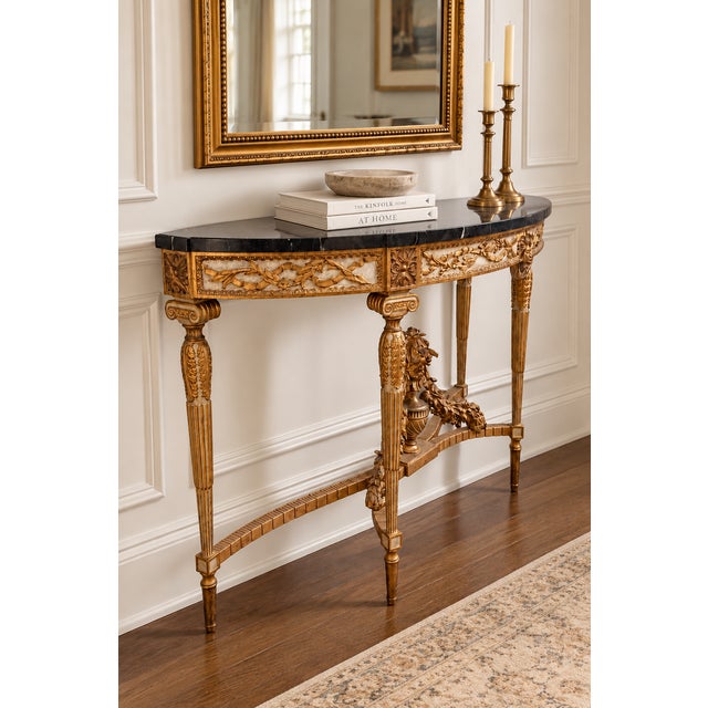An exquisite Louis XVI-style demi-lune console table featuring elaborate giltwood ornamentation and a striking black...