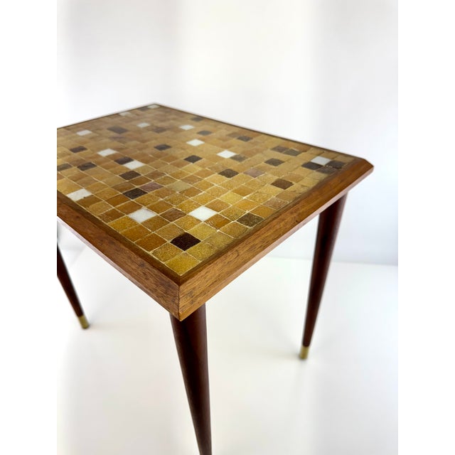 A striking mid-century modern side table featuring a geometric mosaic tile top in warm shades of caramel, amber, cream,...