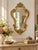 An elegant vintage wall mirror featuring a richly ornamented, Rococo-inspired frame with graceful scrolling curves,...