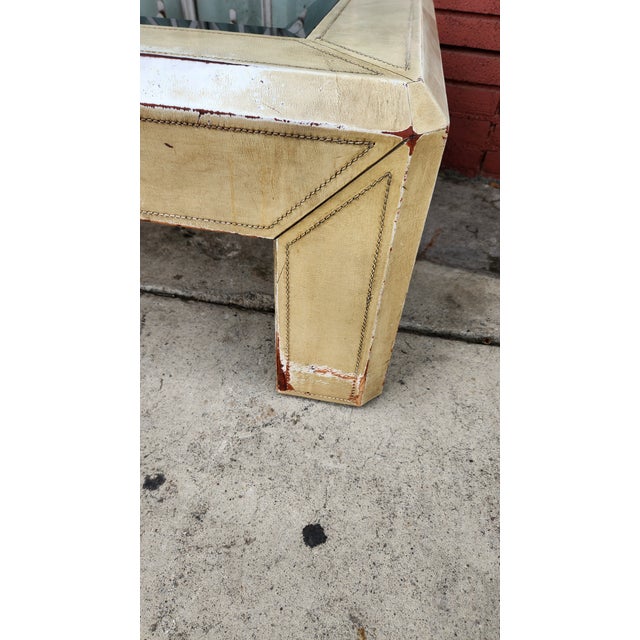Distressed Leather coffee table has a rectangular glass top insert. The frame is covered in a cream-colored material with...