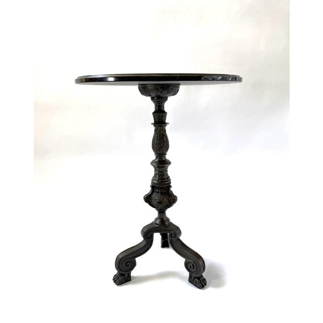 Elegant bronze side table featuring a striking round dark marble top with subtle natural veining. The table rests on a...