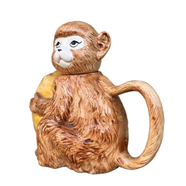 A utterly charming and expertly crafted majolica teapot in the form of a wide-eyed monkey clutching an oversized yellow...