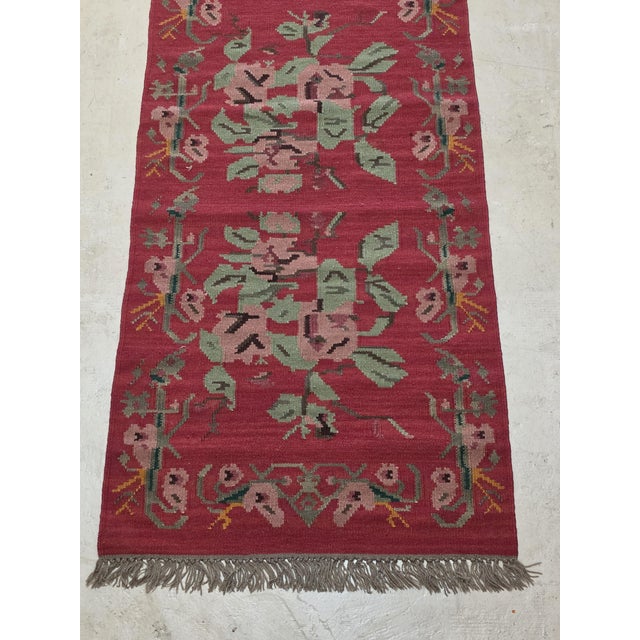 Vintage handwoven kilim runner featuring a rich brick-red background adorned with charming oversized floral motifs in soft...