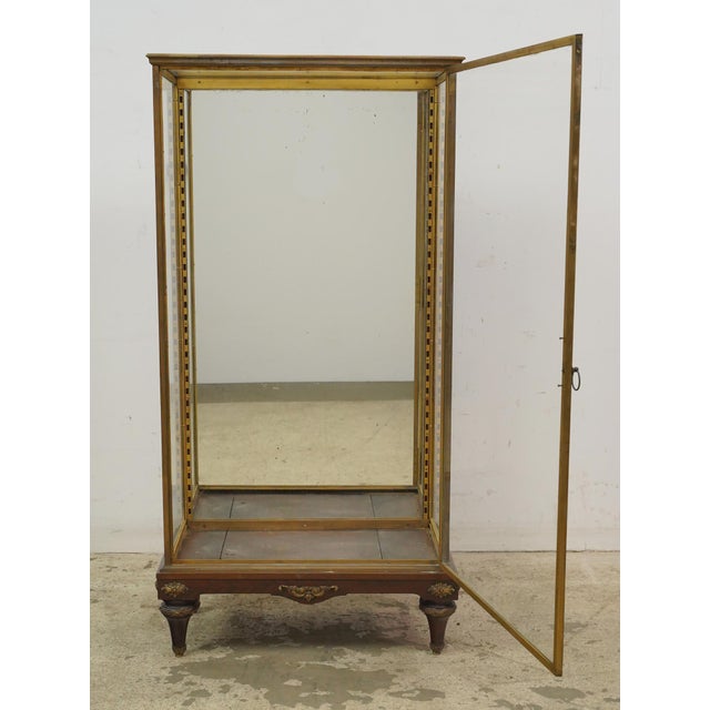 The vitrine features a clear glass door and sides, a mirrored back, and a simple metal ring handle. The bottom of the...