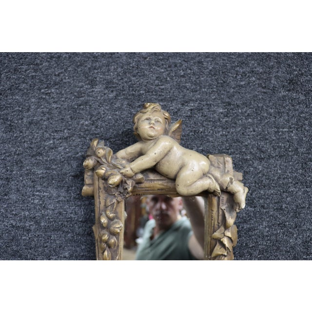 Hand-carved Italian wall mirror in the Baroque style featuring a reclining winged cherub above a rustic frame adorned with...