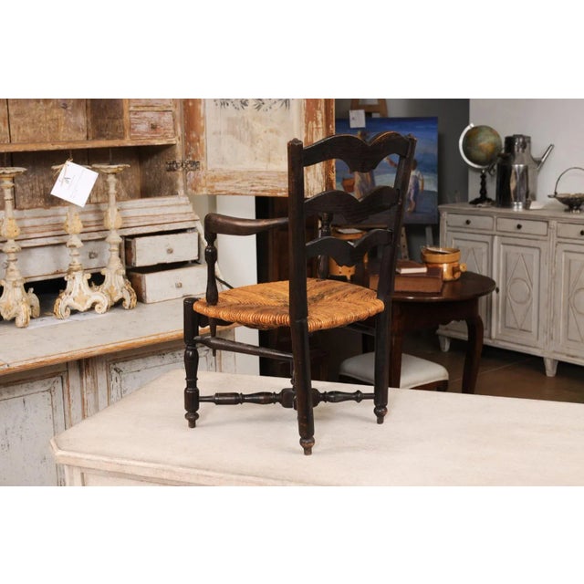 A rustic French dark oak child's chair from the 19th century with carved ladder back, open arms with turned supports, rush...