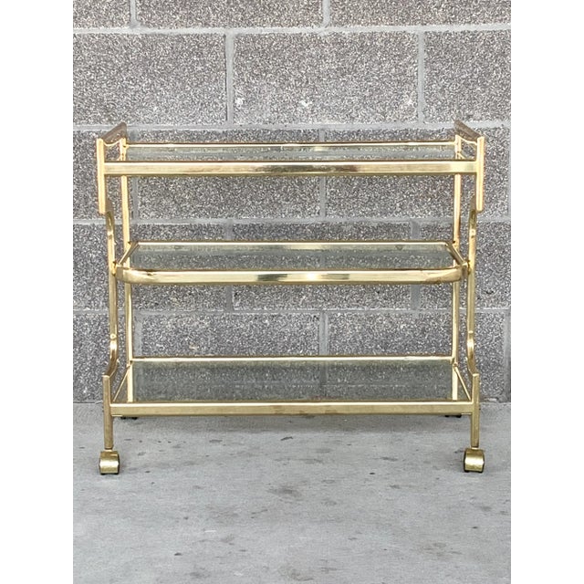 Stunning vintage brass colored bar cart. Milo Baughman style. With a sleek Hollywood regency design. Features three glass...