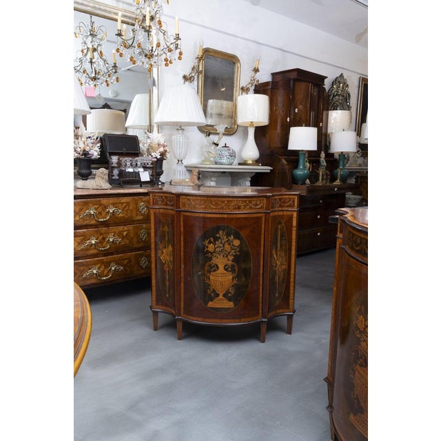 This is one of a pair of elegant English Neoclassical demilune cabinets crafted in richly figured mahogany with satinwood...