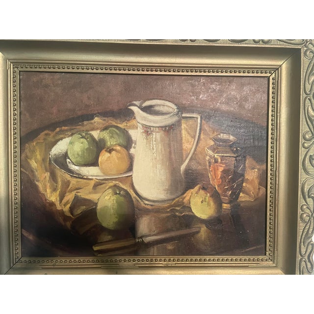 Antique framed still life oil painting featuring a white pitcher, plate of apples, and a decorative jar on a draped yellow...