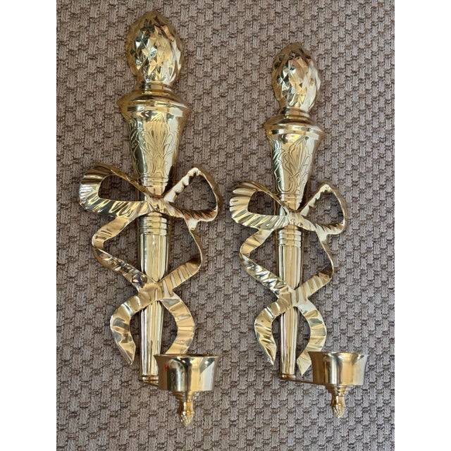 Shiny pair of 1980s solid brass torch and bow wall sconce candle holders. Detailed etchings and topped with a pineapple. I...
