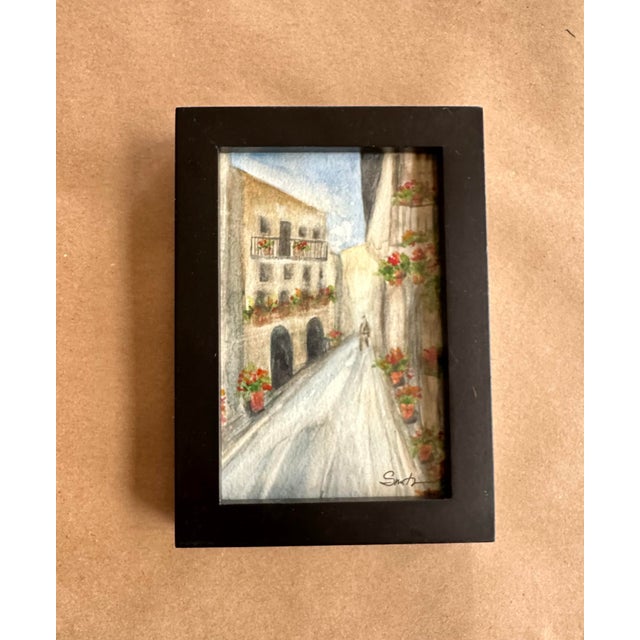 A delicate and romantic watercolor depicting a quiet European-style street lined with warm-toned buildings, flower boxes,...