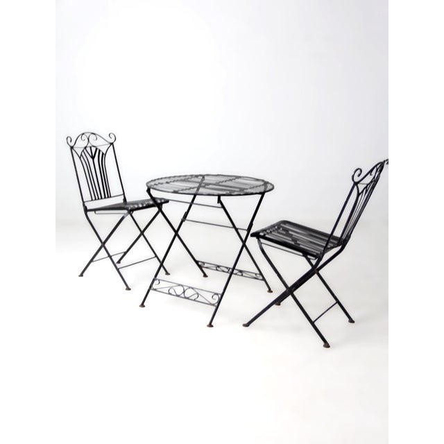 A mid-century wrought iron patio set in the style of classic French bistro seating, featuring graceful scrollwork backs...