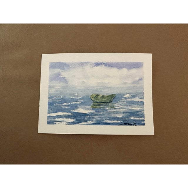 A serene original watercolor depicting a small green boat resting quietly on shimmering blue water beneath a softly...