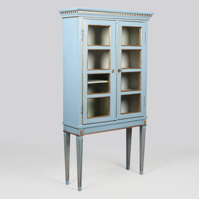 A blue-painted Danish Louis XVI display cabinet. The piece features subtle gold accents and two crossed doors. The...