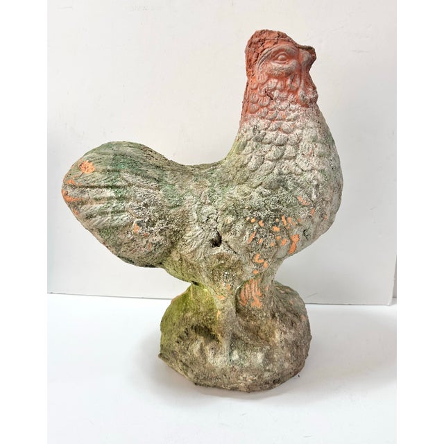 A weathered terracotta rooster garden statue with moss, mineral deposits, and softened surface texture from years...