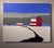 This oil on panel artwork depicts a serene winter landscape featuring a striking red barn and smaller structures set...