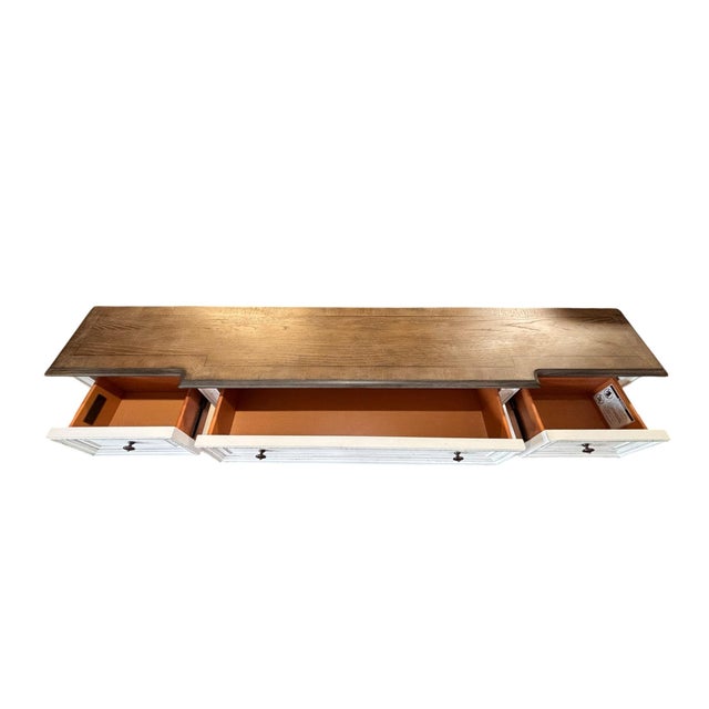 Handsome long console table featuring a warm dark-stained wood top contrasted with a distressed antique-white painted...