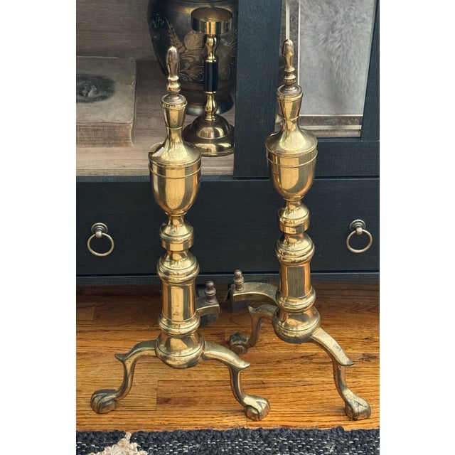 A 19th-century pair of brass andirons in classical architectural form with turned stems and claw feet, showing a warm,...