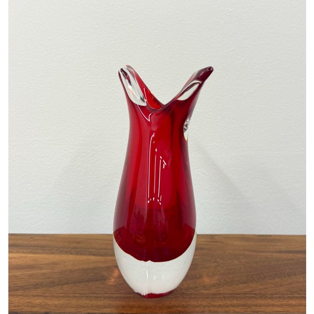 A sculptural Murano-style art glass vase featuring a vivid ruby-red interior encased in thick clear glass. The substantial...