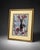 Framed floral still life artwork featuring flowers, fruit, glassware, and a table setting in a classic decorative style....