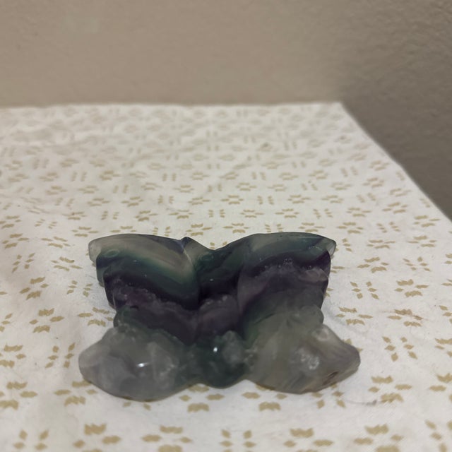 This hand-carved butterfly is formed from rainbow fluorite, displaying soft bands of purple, green, and translucent tones...