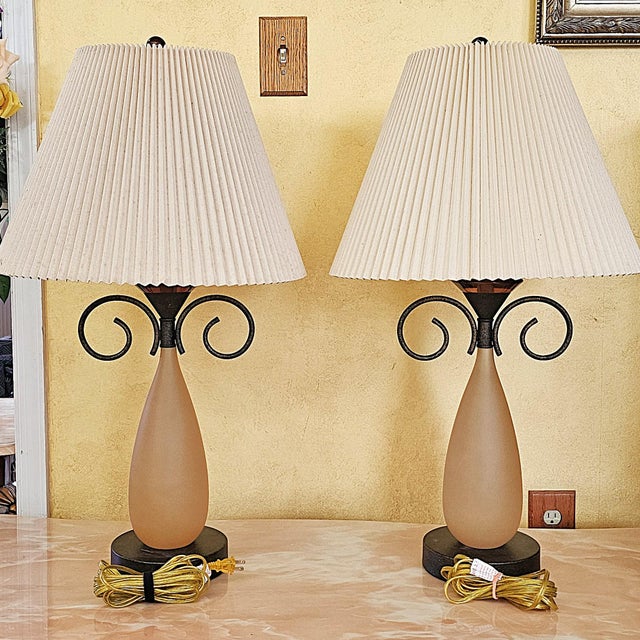 A sculptural pair of table lamps featuring elongated teardrop frosted glass bodies in a warm amber-orange tone, accented...