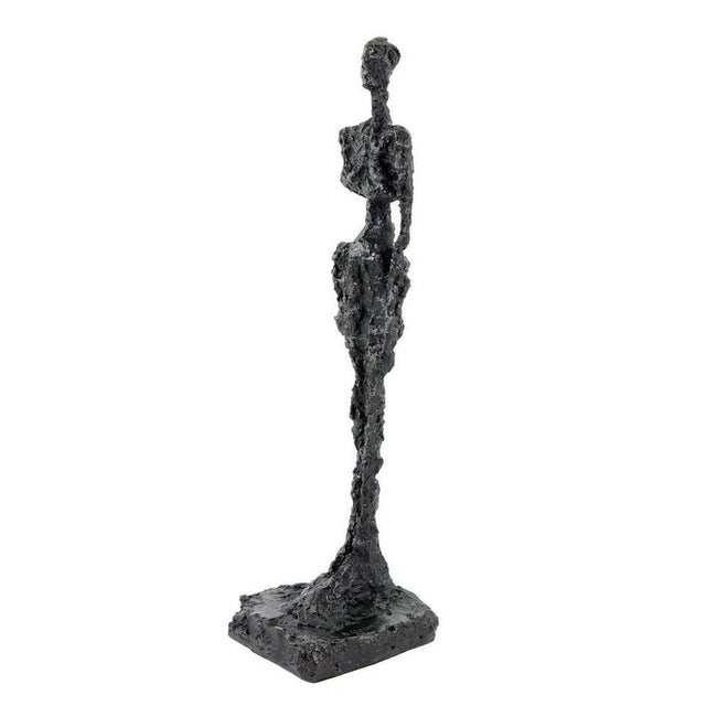 This modern bronze sculpture represents a standing female figure rendered in an elongated and abstract form. Cast in dark...