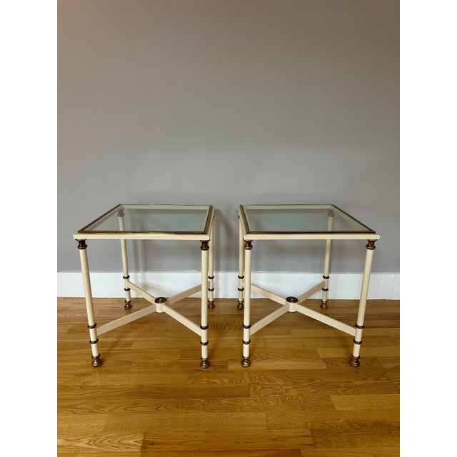 A pair of French side tables in cream lacquer and brass with clear glass tops. The up rights, cross frame support in...