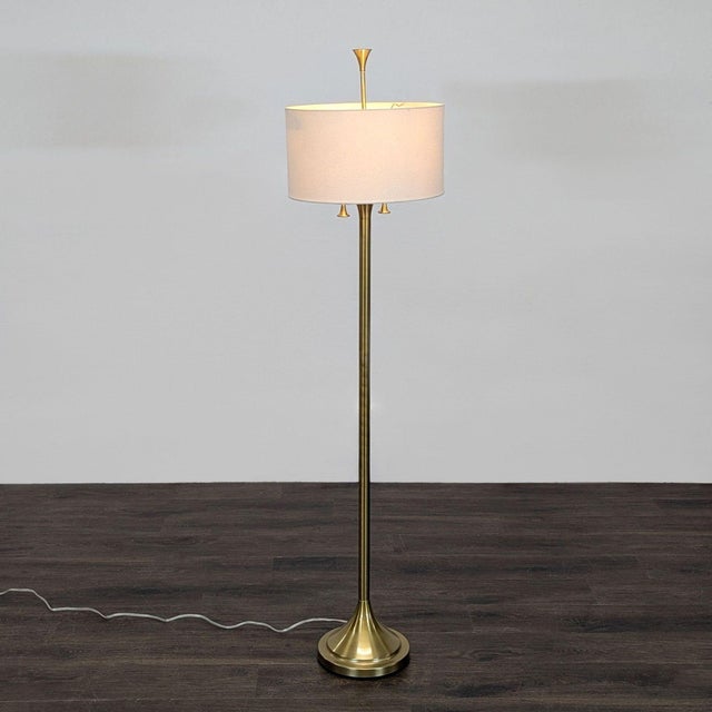 This elegant floor lamp features a sturdy brass base and pole, topped with a simple white fabric drum shade. It includes...