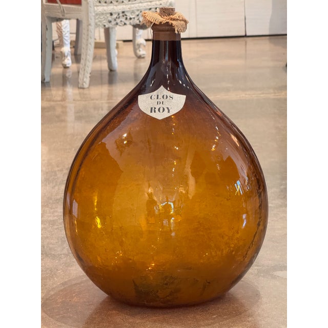 A blown amber glass demijohn with a shield reading "Clos du Roy." Cork stopper with burlap collar.