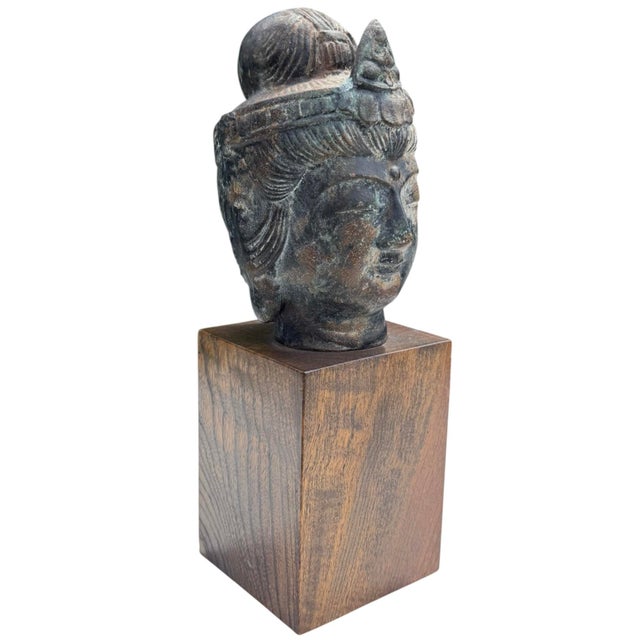 Vintage sculptural Buddha head with a dark, aged bronze finish, mounted on a simple solid wood pedestal. The serene...