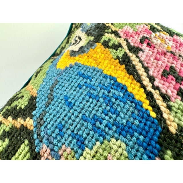 A richly detailed needlepoint pillow featuring a vibrant parrot perched among lush greenery and pink blossoms. The...