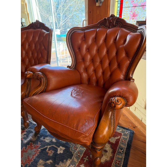 A handsome pair of tufted leather armchairs featuring richly carved wood frames and classic button-tufted backs....