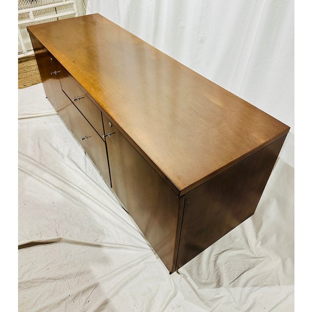 A sleek and understated Mid-Century Modern credenza featuring clean architectural lines, warm walnut-toned casework, and...