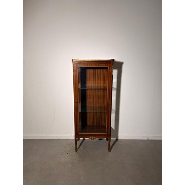 An elegant display cabinet featuring a warm brown wood finish, glazed front and sides, and decorative gold-tone metal...