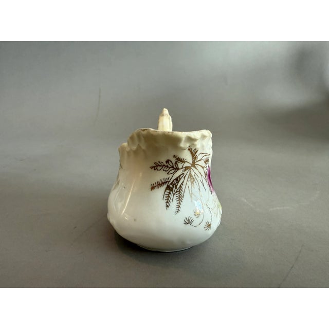 This antique creamer is made of porcelain and features a transfer ware design with purple and white flowers accented with...