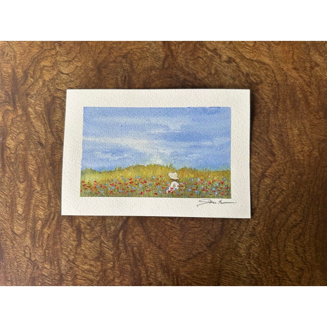 A charming and whimsical watercolor painting featuring a field of delicate wildflowers beneath a soft blue sky. A little...