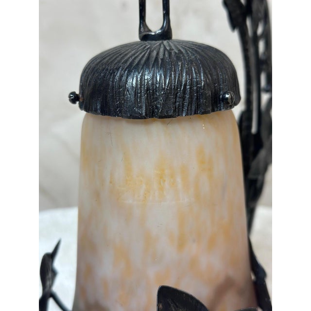 A charming Art Deco lamp combining a wrought iron frame with a glass diffuser in shades of yellow and amber, bearing the...