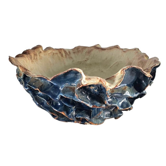 A bowl as sculpture, its ruffled edge folding like waves caught mid-motion. A deep blue glaze, dark and lustrous, clings...